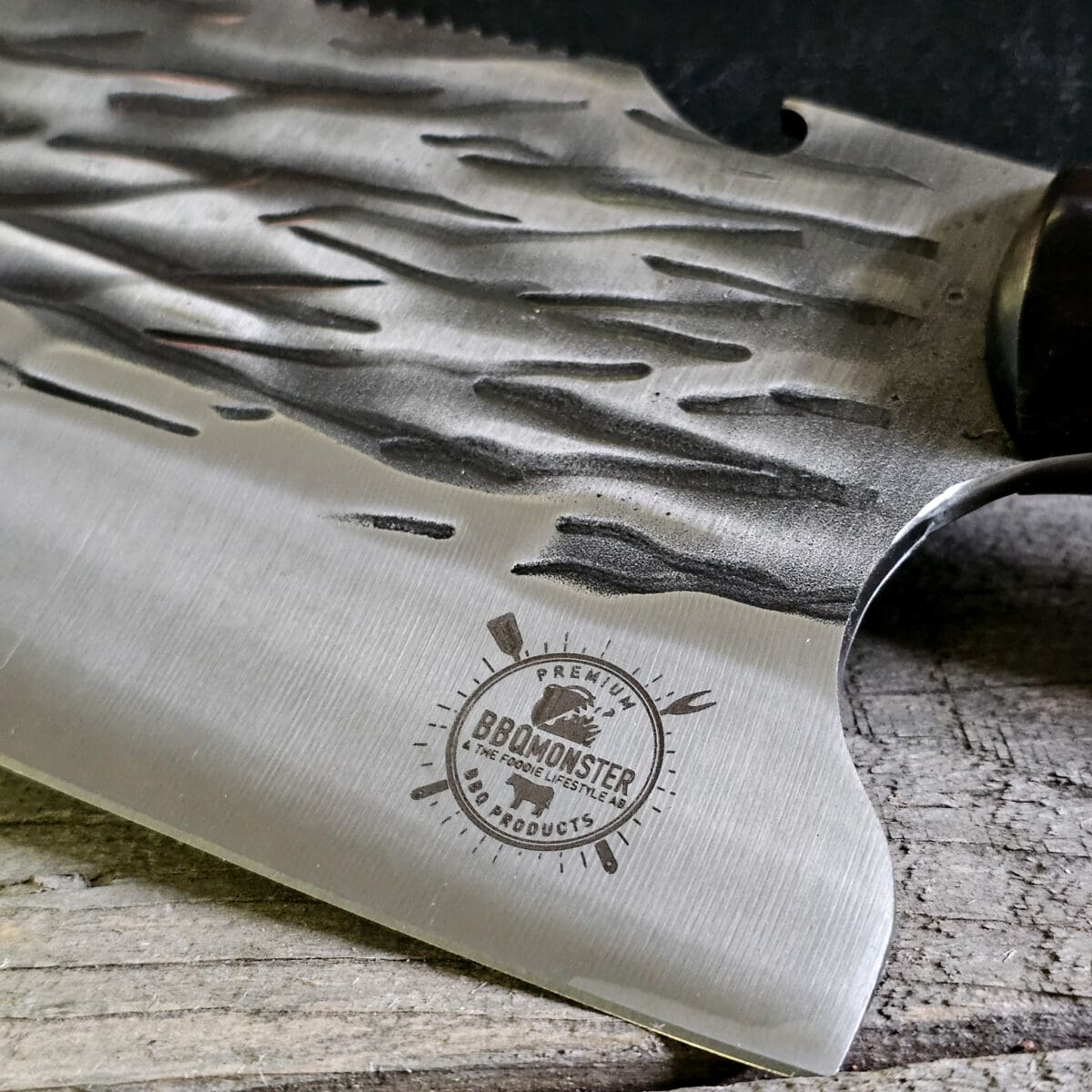 Kökskniv / Cleaver "Fire #7" 29cm - grovt kolstål med fina detaljer och galet vass - med ölöppnare på ovansidan av bladet - säljs för det tredubbla på andra sidor! Söker du en serie av välarbetade och lättskötta kockknivar? Ta en kik på BBQmonsters egna serie knivar! Samtliga knivar i serien kommer i en svart presentförpackning med BBQmonsters sigill "Make BBQ great again" tryckt på locket. Knivarna från BBQmonster har även BBQmonsters logo ingraverad på bladet. Vi sätter bara vår logo på prylar som vi verkligen tycker om - detta är en sådan. Introduktionspris - passa på! En perfekt till grillnörden, en present till pappa på farsdag eller till dig själv helt enkelt! Denna modell av kökskniv är av typen som på många sidor benämns som en "serbisk kockkniv". Hur väl den beskrivningen stämmer låter vi var osagt men det man kan säga om denna kniv är att den perfekt när du skall skapa snittytor som är jämntjocka, detta då bladet är imponerande 10cm högt.  Att typen av kniv blivit populär beror sannolikt på den smidiga hantering att plocka upp hackad lök, koriander och annat med ETT svep - bladet är som "skyffel". Bladet på denna Fire #7 är grovt, tungt, stabilt, lättskött och bladet har en härlig yta som påminner om just "fire" - lågor. Tillsammans allts som allt gör detta kniven rejält "intstagramvänlig" och photogenique. På ovansidan av bladet sitter en ölöppnare så du slipper slita på snusdosan. (Vill du hellre ha en ölöppnare uppskruvad på väggen så ta en kik på denna Bulldog i gjutjärn!) Bladet är genomgående och avslutas med handtaget som är av polerat och lackat ädelträ och tre nitar av mässing. Skaftet ger ett skönt grepp och är enkelt att rengöra. Handtaget passar både höger- och vänsterhänta.  Kolstål med blandning X50CrMoV15 Hårdhet 56° HRC vilket är ett relativt mjukt stål - något som gör att du själv enkelt kan hålla din kniv vass. Total längd: 29 cm Längd på bladet: 17 cm Längd på skaftet: 12 cm